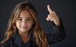 A young girl with long, wavy hair smiles warmly as she gives a thumbs up gesture. She is wearing a smart black blazer, and the dark background behind her 