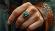 A close-up view of a person's hand displaying an ornate ring with a large emerald gemstone against a textured fabric background. 