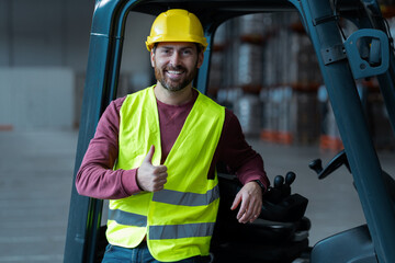 Wall Mural - Bearded man, driver showing thumbs up, wearing helmet, standing near forklift, looking at camera