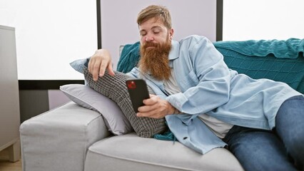 Wall Mural - Serious young irish guy, redhead with a beard, sitting on the sofa in his apartment, looking upset while texting on his smartphone indoors