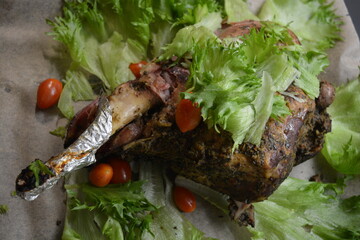 Wall Mural - Eating, cooking delicious meat, sheep leg on the bone marinated and baked in the oven. A large piece of lamb arranged on brown parchment paper with green lettuce leaves and small cherry tomatoes.