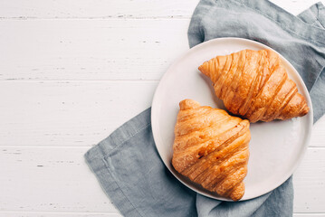 Plate with two fresh croissants on white wooden table. Top view.