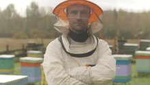Tilt Up Medium Slowmo Portrait Of Caucasian Male Beekeeper In Protective Clothing Posing With Hands Folded At Apiary With Colorful Wooden Bee Hives In Background