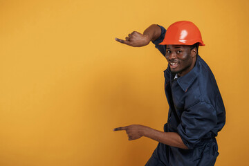 Wall Mural - Guy in hard hat showing copy space by fingers. Handsome black man is in the studio against yellow background