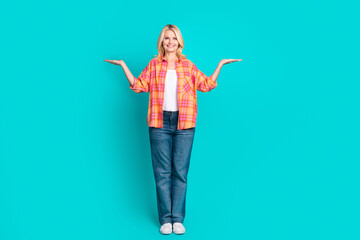 Full length photo of cheerful woman dressed plaid shirt denim pants comparing products empty space isolated on turquoise color background