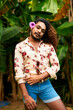 Confident gay man with a bright flower in his curly hair stands outdoors. Short blue shorts, floral shirt, hand on hip, sultry gaze in a tropical setting. Expresses pride, freedom, and diversity.