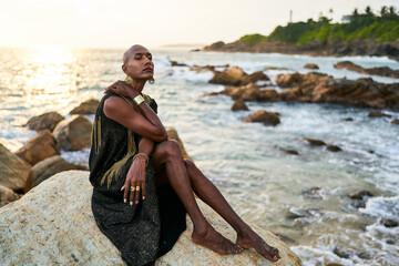 Wall Mural - Non-binary black person in luxury dress, golden jewelry on beach rocks in ocean. Trans ethnic fashion model wearing jewellery in posh gown poses gracefully in tropical seaside location on sunset.