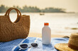 Bottle of sunscreen on striped towel next to wicker bag, straw hat, and sunglasses on sunny beach, with ocean horizon in background. Essential items for skin protection, leisure day by sea.