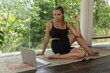 A young caucasian athletic pretty woman doing a warm-up before training on a mat looking at the laptop screen at home with panoramic windows overlooking green trees. A girl doing stretching