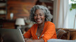 A happy cheerful African-American woman in her 60s with a laptop sits at her desk, smiling, looking at the screen, writing something down, making a shopping list	