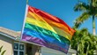Rainbow pride flag waving in front of a house with palm trees.