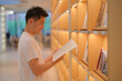 handsome Asian young male student hold and read book at bookshelf in college library. side view
