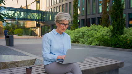 Mature woman working online typing email business chat in internet using laptop pc computer senior old businesswoman in city modern urban street downtown female entrepreneur work with project outside