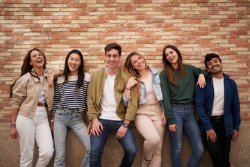 Portrait group of international cheerful friends posing together hugging looking smiling at camera. Happy generation z people leaning on brick wall outdoor. Relationships, friendship and community