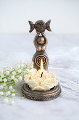 Sticker - amulet of Triple Goddess figurine, floral candle and lilies of the valley flowers close up, abstract light background. Symbol of the Triune Moon. esoteric ritual for Imbolc, Ostara. witchcraft