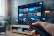 © PAOLO - A hand holding a remote control in front of a smart TV with streaming services on the screen, in a close up shot.