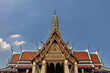 Temple at Grand Palace (Wat Phra Kawe) in Bangkok, Thailand. Ornate gold decorations; green, red & orange tiled roof. Decorated prang in background. Blue sky and clouds behind. 
