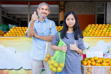 Wall Mural - portrait teen girl and senior grandfather carry net tole bag of oranges and doing thumbs up in front of community fruits store for support using environmentally friendly bags