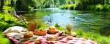 Fototapeta  - Summer picnic with a basket of bread, pastries, and fruits on a red checkered cloth next river.