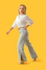 Wall Mural - Stylish young woman in jeans walking on yellow background