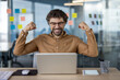 Joyous male professional in glasses celebrating a successful deal or achievement while working on his laptop in a modern office setting.