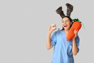 Sticker - Happy female dentist in bunny ears with toy carrot and jaw model on grey background. Easter celebration