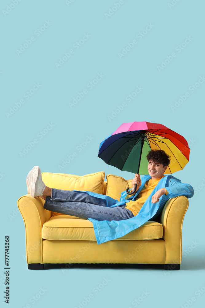 Young man with umbrella lying on sofa against blue background