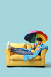 © Pixel-Shot - Young man with umbrella lying on sofa against blue background