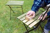 Fototapeta Tulipany - Man is cleaning and restoring wooden outdoor furniture, sanding the weathered wood to remove algae before oiling or painting for a neat garden season, copy space