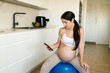 © Ezequiel Giménez/Stocksy - Pregnant woman on fitness ball using cellphone at home