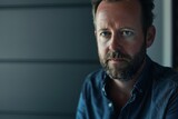 Fototapeta  - Pensive mature man in blue shirt, contemplative gaze, dimly lit room, personal challenges, life decisions, intimate portrait.

