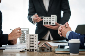 A professional businessman in a suit conducts a meeting at a desk while showcasing a model of a condominium or apartment building, discussing real estate development plans.