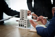 © Ratirath - A professional businessman in a suit conducts a meeting at a desk while showcasing a model of a condominium or apartment building, discussing real estate development plans.