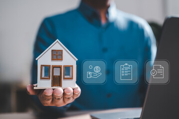 real estate, conceptual, contract, estate, insurance, mortgage, ownership, residence, property, apartment. A man is holding a small house model in his hand while looking at a laptop.