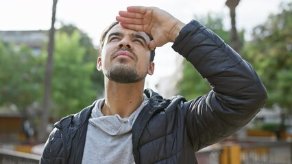 Wall Mural - A young hispanic man in a jacket stands in a park, looking upwards shielding his eyes from the sun.