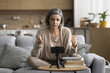 © fizkes - Middle aged woman sitting on sofa in living room records new videoblog, speak at cellphone camera fixed on tripod, share experience, talk to internet audience. Online communication, coaching, blogging