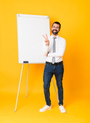 Wall Mural - Full-length shot of businessman giving a presentation on white board over isolated yellow background smiling and showing victory sign