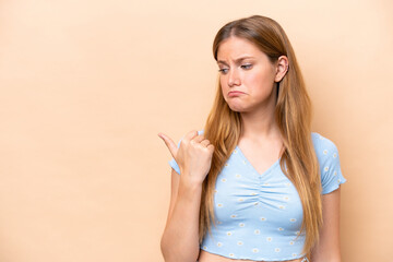 Wall Mural - Young caucasian woman isolated on beige background unhappy and pointing to the side