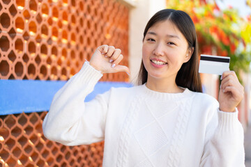 Wall Mural - Pretty Chinese woman holding a credit card at outdoors proud and self-satisfied