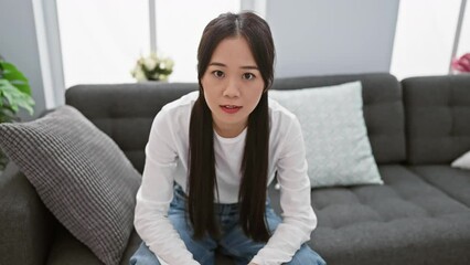 Canvas Print - A young asian woman sitting thoughtfully in a modern living room with a grey sofa and plant decoration.