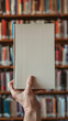 © Irina B - A man's hand holds a book with a blank white cover against the background of a blurred library or bookstore. Mockup, template. Day of Knowledge. World Book and Copyright Day