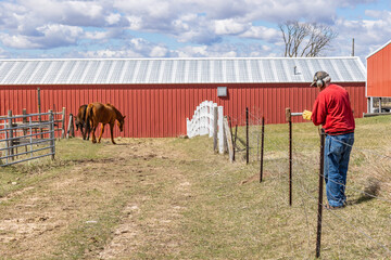 A man pounding in a fence post with a sledgehammer in a farm lane with two horses and a barn in the background. 