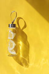 Wall Mural - Glass bottle with lemon water drink detox at sunlight on yellow background, copy space. Healthy infused water boost metabolism and weight loss. Aesthetic still life minimal, summer drink