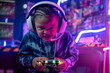 African child boy wear headphone excited playing video game, controlling characters with joystick at colorful neon light living room at night, E-sport streaming game online