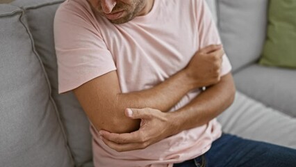 Sticker - A young man in pain holding his elbow while sitting on a couch indoors, suggesting injury or discomfort.