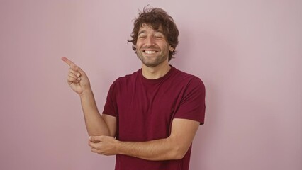 Wall Mural - Cheerful young hispanic man in a t-shirt confidently pointing to the side with a friendly smile, isolated over a pink background