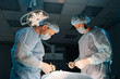 © dikushin - Low-angle view of surgery professionals operating patient. Male doctors using metal tools at work. Round lamps illuminating surgery dark room. Medical team performing surgery. Concept of emergency.