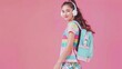 Full length portrait of young cheerful girl in summer wear with backpack, listening to music in white headphones, looking at camera, over pink background