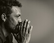 A man with a beard and a green shirt is praying. He is looking down and his hands are clasped together