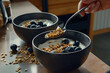 Close up of a spoon holding cereal in low-fat yogurt and blueberries above the black ceramic bowl on the kitchen counter, another bowl with only blueberries in, next to the first one. The other hand h